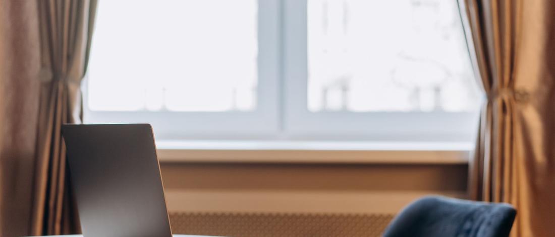 Modern laptop sits on a wooden table with curtains and window in the background.
