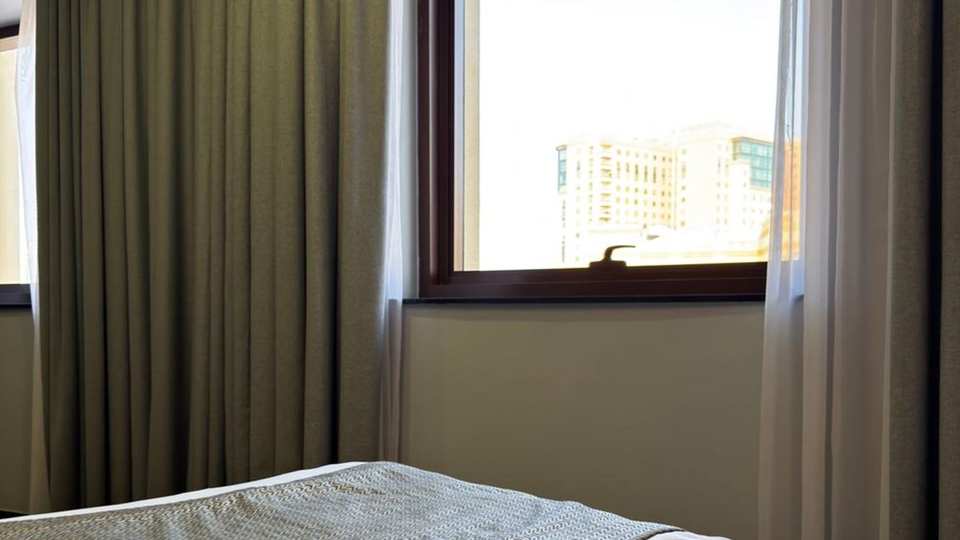 Modern hotel room with bed and cityscape view through a large window.