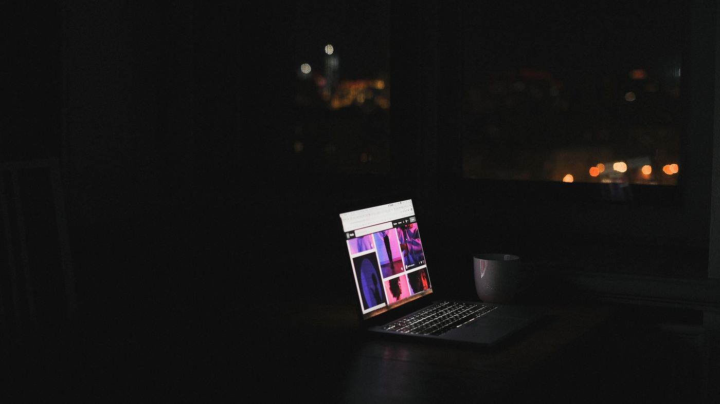A laptop glows in a dark room at night with a cityscape through the window. Ideal for tech and solitude themes.