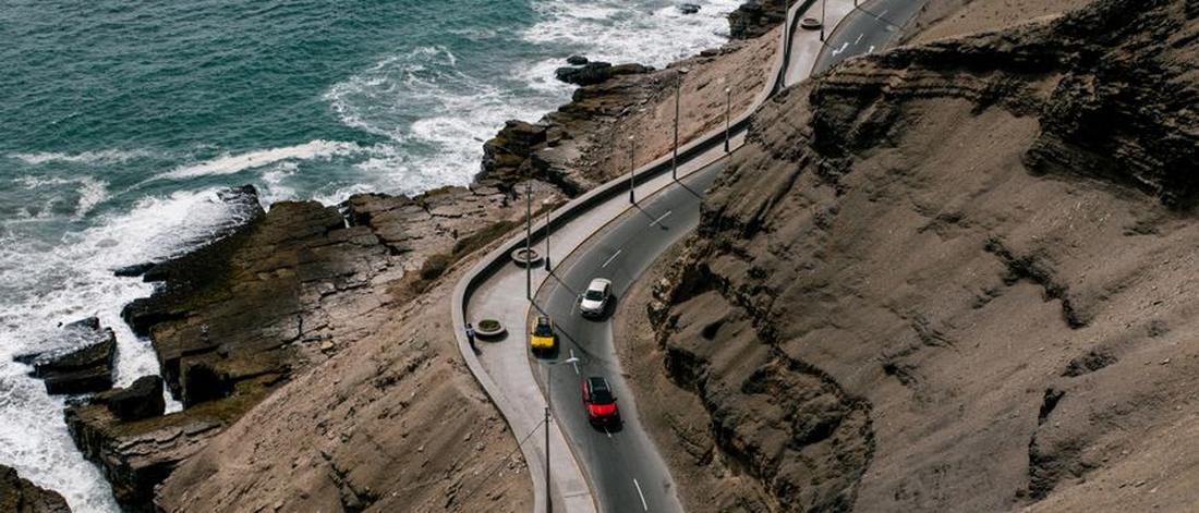 From above of various cars driving on curvy road going on rough rocky hill near stormy ocean