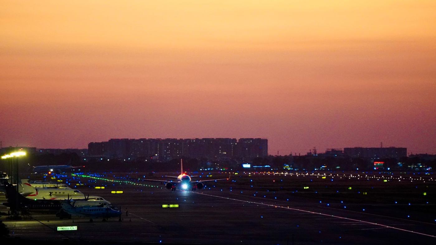 Airplane preparing for takeoff on an illuminated runway, against a sunset cityscape backdrop.